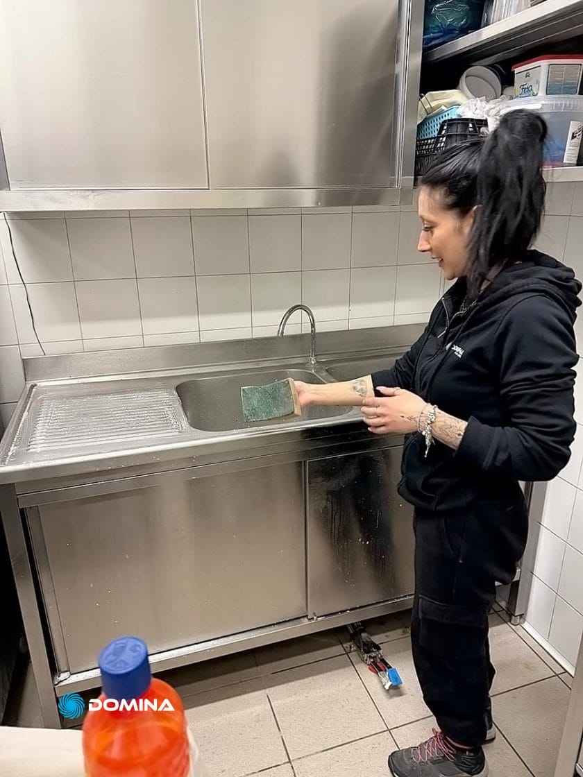 Una persona lava diligentemente i piatti in un lavandino in acciaio inox, tenendo in mano una spugna. Sopra il lavandino si possono vedere scaffali con le forniture essenziali, un'organizzazione efficiente che ricorda un'impresa pulizie per ristoranti Milano.