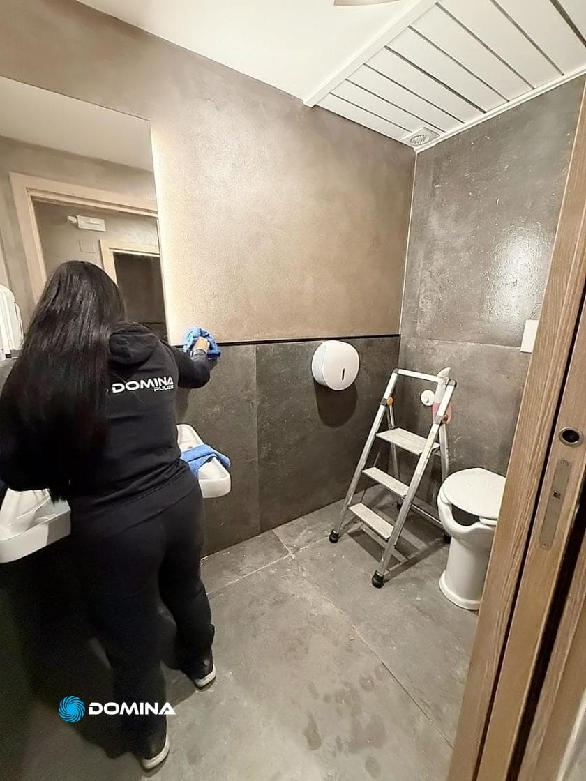 Una persona di un'impresa di pulizie per ristoranti di Milano pulisce il lavandino di un bagno. Nelle vicinanze, una scala è vicina al water e un distributore a parete contrasta con le pareti marroni e grigie.