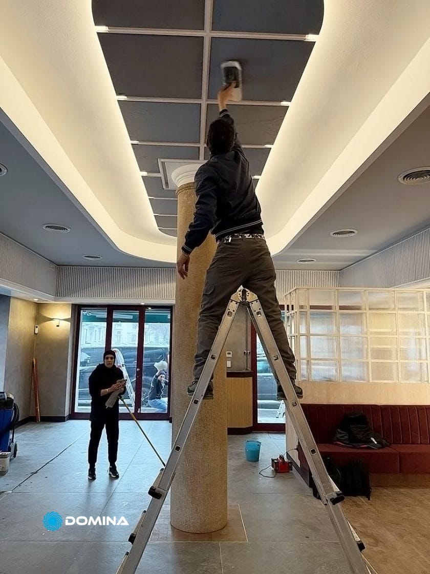 In una stanza con arredamento moderno, qualcuno sale su una scala e pulisce il soffitto mentre un altro spazza il pavimento lì vicino. Questa scena riflette gli standard meticolosi di un'impresa di pulizie per ristoranti Milano, che assicura che ogni angolo brilli.
