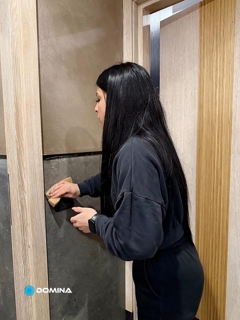 Una persona con lunghi capelli neri e un abito scuro leviga una superficie di legno con la carta vetrata in un ambiente chiuso, proprio come fa un'impresa di pulizie per ristoranti di Milano per garantire che ogni dettaglio brilli in modo impeccabile.