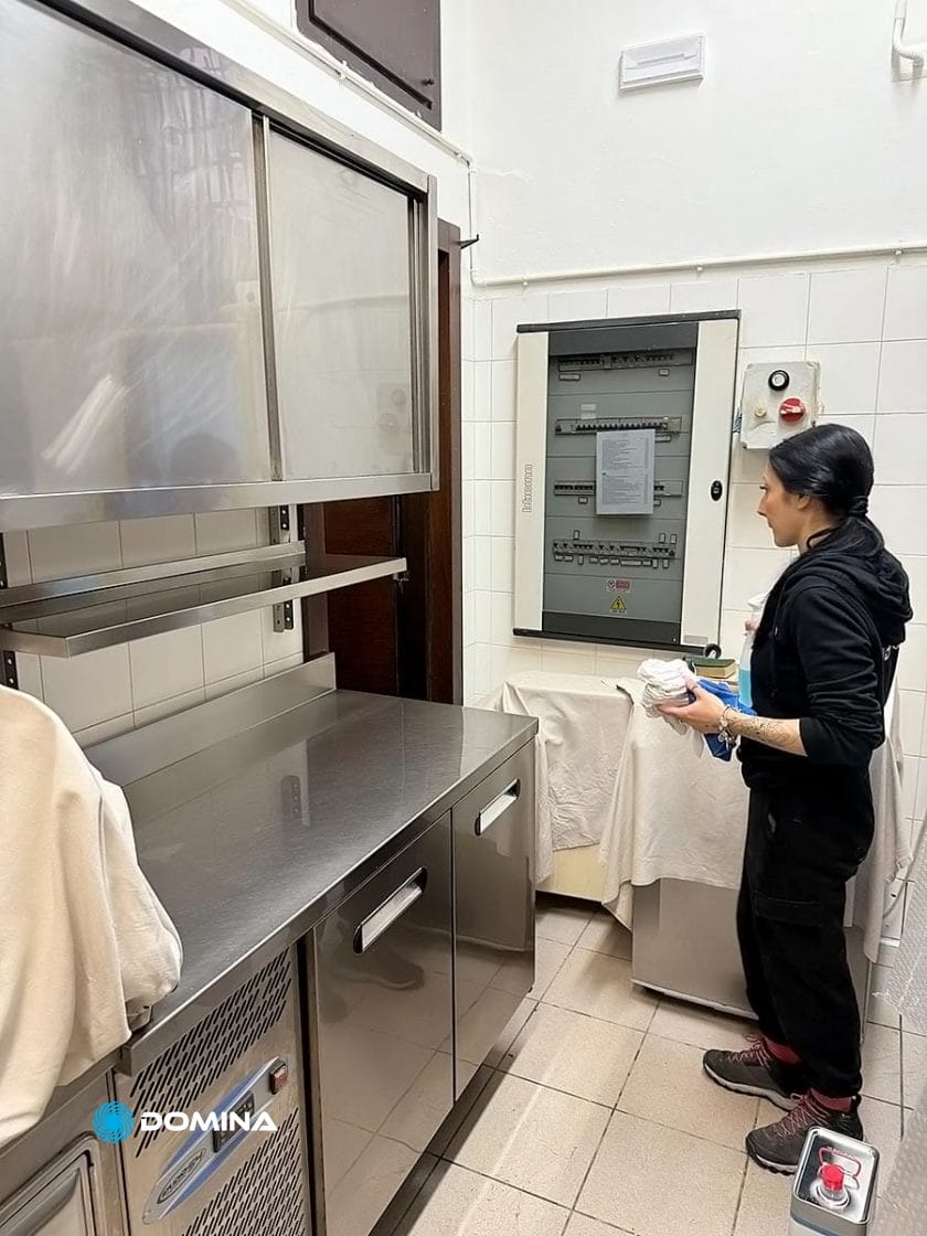 Una persona è in piedi in una cucina accanto a un bancone e a un elettrodomestico in acciaio inossidabile, tenendo in mano degli oggetti. Sullo sfondo, una macchina coperta da un asciugamano allude agli standard meticolosi mantenuti da impresa pulizie per ristoranti Milano.