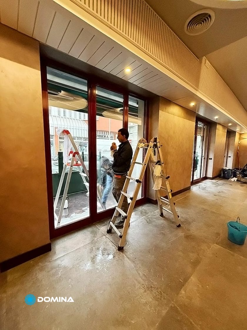 Una persona è in piedi su una scala all'interno di un edificio, che sbircia attraverso grandi porte di vetro. Un'altra scala è visibile all'esterno, a suggerire i lavori in corso. Il pavimento sembra incompiuto e un secchio blu si trova più in basso, forse parte degli sforzi di un'impresa di pulizie per ristoranti Milano.