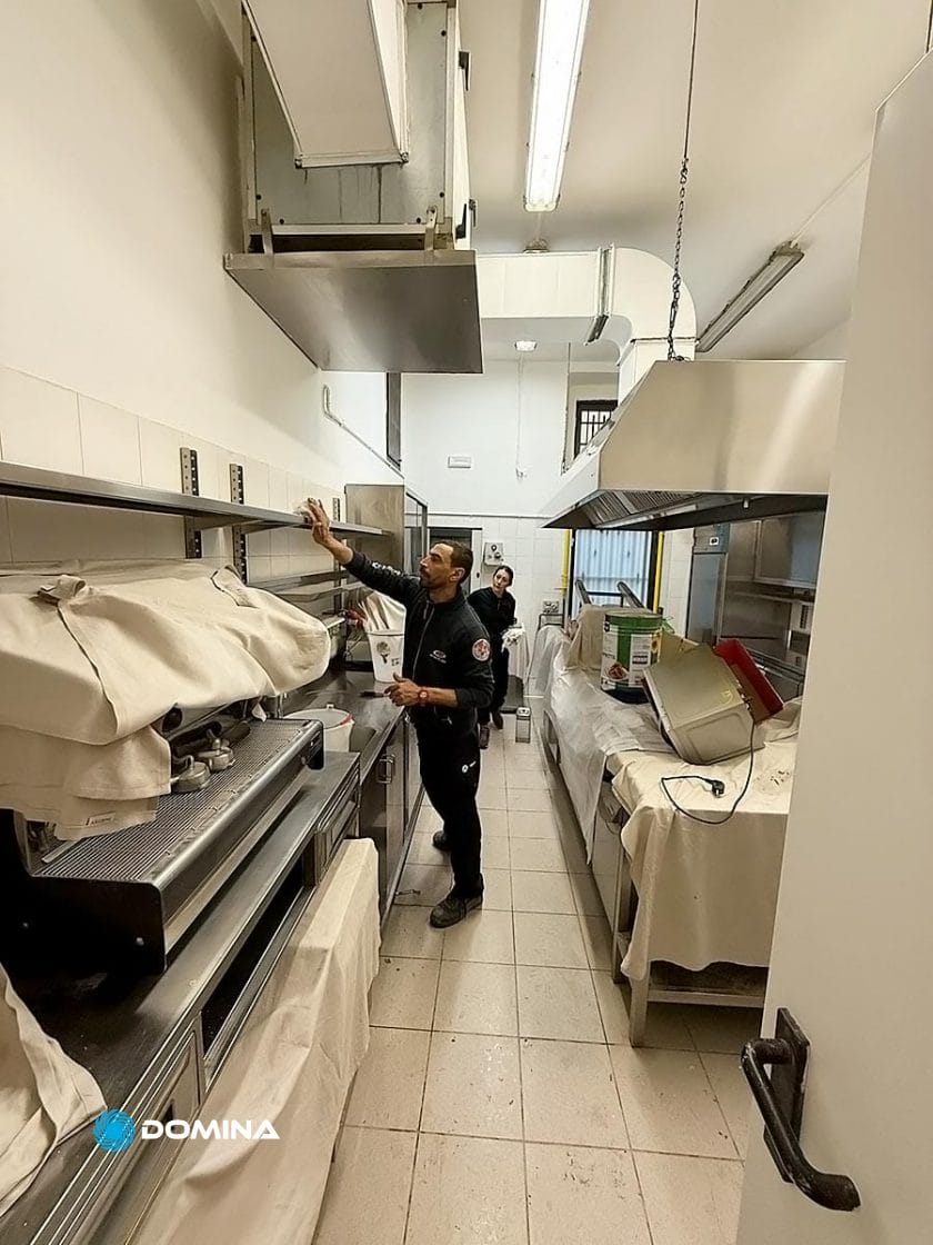 Due persone ispezionano una cucina commerciale, le cui superfici in acciaio inossidabile brillano sotto una forte illuminazione fluorescente, che ricorda gli standard delle migliori imprese di pulizie per ristoranti di Milano, con elettrodomestici coperti che garantiscono la massima igiene.