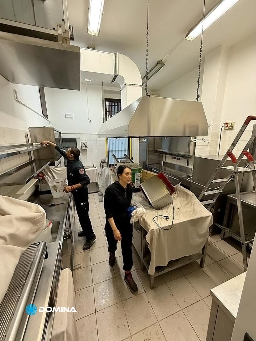 In una cucina commerciale in fermento, due persone lavorano insieme in modo efficiente, lei con un panno in mano e lui che manovra una rastrelliera, mettendo in mostra la competenza di impresa pulizie per ristoranti Milano. Elettrodomestici in acciaio inossidabile scintillanti e attrezzature ben coperte completano la scena.