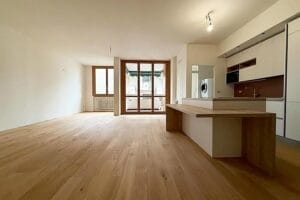 Ampia stanza vuota con pavimenti in legno, una moderna zona cucina perfetta per le pulizie di casa, primo ingresso e grandi finestre che conducono a un balcone con vista sulla bellezza di Milano.