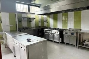 Cucina commerciale con superfici in acciaio inox, una grande stufa, forno e ripiani portaoggetti. Pareti piastrellate di verde e bianco evocano lo spirito del Ristorante Milano Centro, mentre un'alta finestra con sbarre mette in mostra l'attenzione ai dettagli che si riscontra negli ambienti del Servizio Professionale.