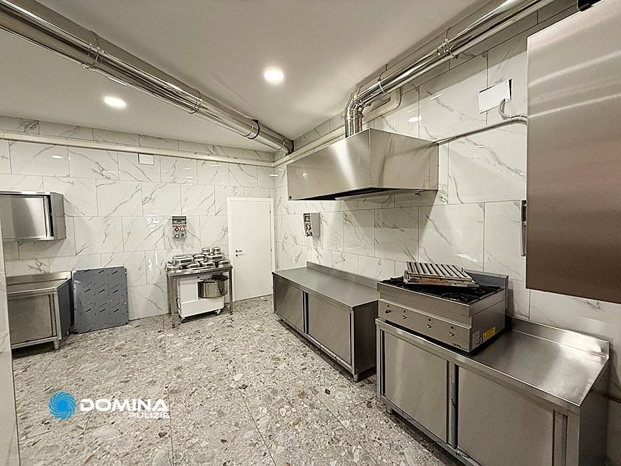 Una cucina commerciale con elettrodomestici in acciaio inox, pareti in marmo e pavimento in pietra vanta una pulizia di livello professionale. Con attrezzature come forno, grill e tavoli in metallo sotto una luce intensa accanto a tubi a soffitto visibili, è adatta sia a locali che a ristoranti.