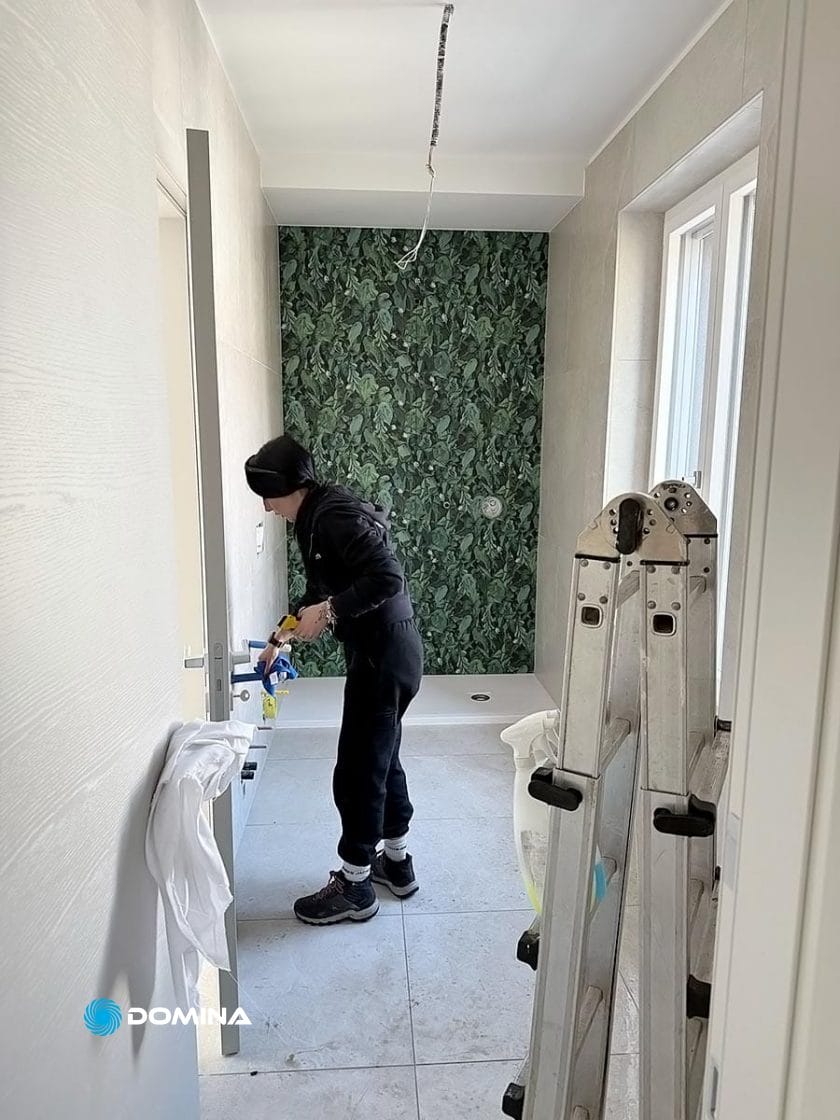 Una persona in abiti neri lavora alla ristrutturazione di un bagno per Domina, un'impresa di pulizie di Nova Milanese, tenendo in mano degli attrezzi vicino a una porta parzialmente aperta. In primo piano si trova una scala a pioli ripiegata; la parete della doccia è ricoperta da carta da parati con motivi a foglie verdi.