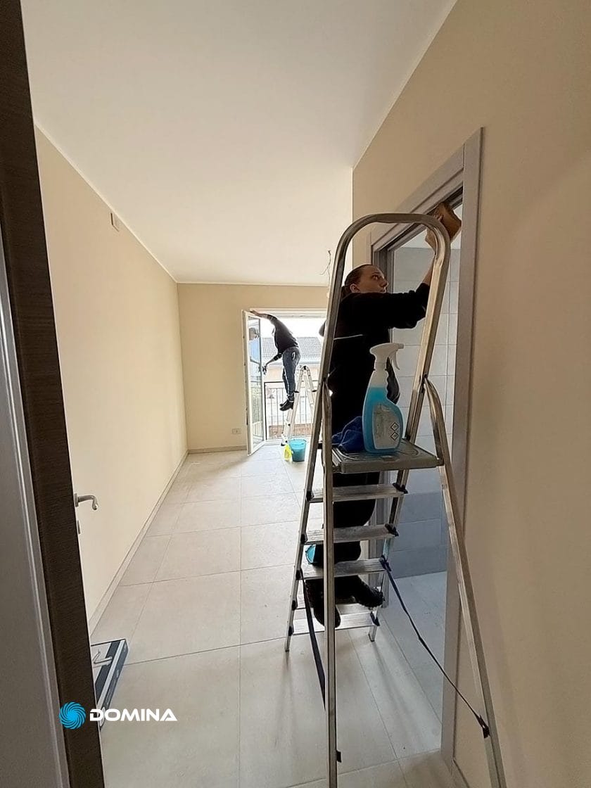 Due persone sono impegnate in pulizie di fine cantiere, pulendo meticolosamente le finestre di una stanza. Una persona sta comodamente in piedi su una scala mentre l'altra lavora fuori sul balcone, circondata da una serie di prodotti per la pulizia.