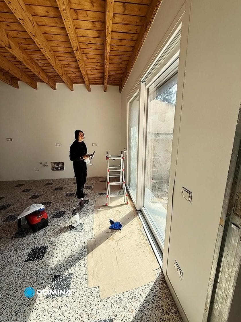 Una persona è in piedi in una stanza soleggiata con pavimenti piastrellati, vicino a una grande porta a vetri, con un attrezzo in mano. Una scala, prodotti per la pulizia della Domina Impresa di Pulizie di Nova Milanese e del cartone sono sistemati vicino alla porta.