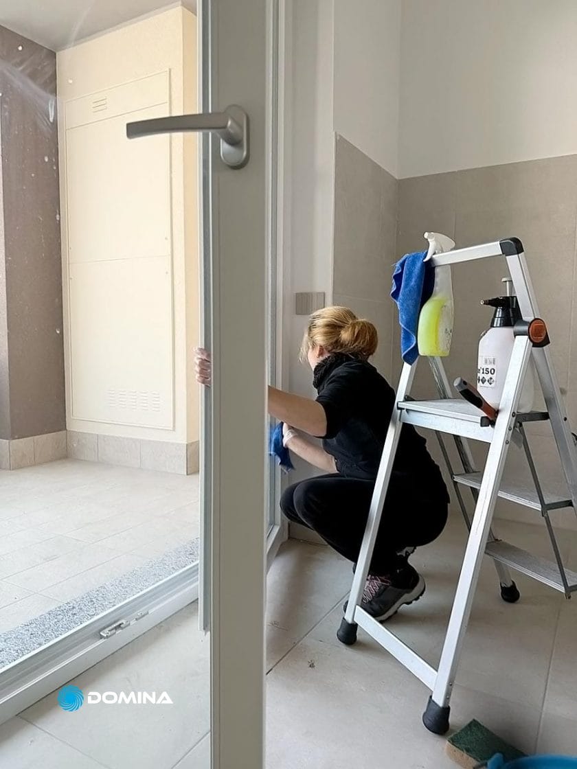 Una persona sta pulendo comodamente una porta a vetri usando un panno e uno spruzzatore, dimostrando abilità simili a quelle di Pulizie di Fine Cantiere. Una scala a pioli con prodotti per la pulizia extra è lì vicino, per far sì che ogni angolo delle case di Paderno Dugnano brilli.