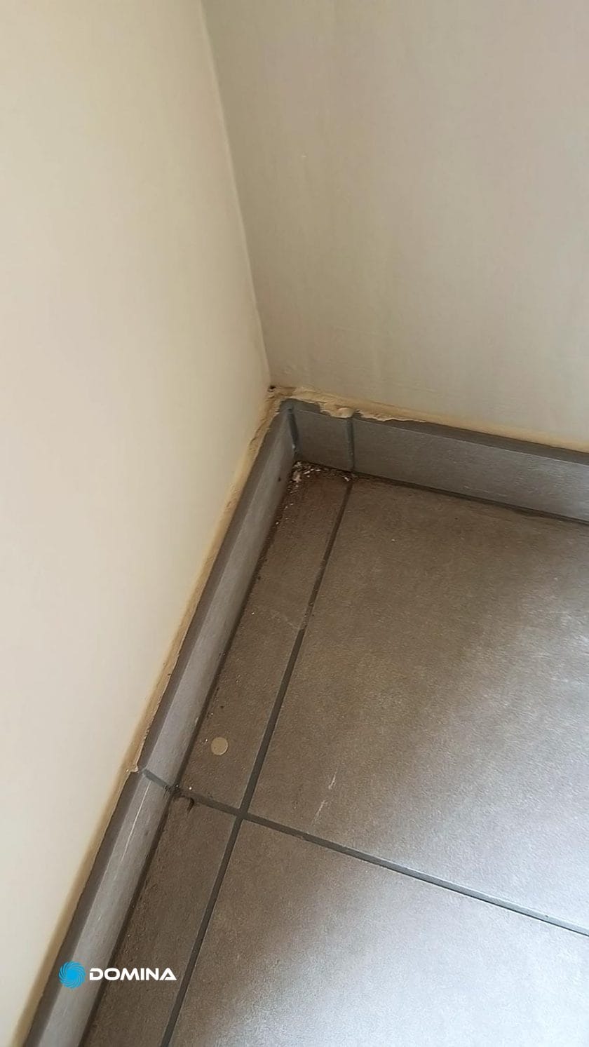 Un angolo di una stanza con pavimento piastrellato e una parete beige a Paderno Dugnano mostra danni d'acqua visibili e scolorimento lungo la base. Affidati a "Trasformiamo il Caos in Comfort" per portare competenza e precisione nel riportare lo spazio al suo antico splendore.