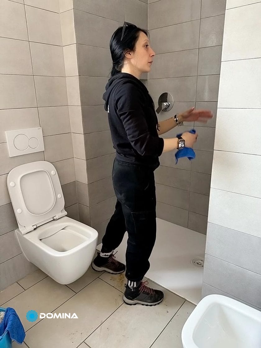 Una persona vestita di nero e stivali è in piedi sotto la doccia con uno straccio per la pulizia, di fronte a una parete piastrellata. Nelle vicinanze, si vedono un water e un asciugamano blu. È come se facessero parte delle "Pulizie di Fine Cantiere", che trasformano il caos in comfort anche qui a Paderno Dugnano.