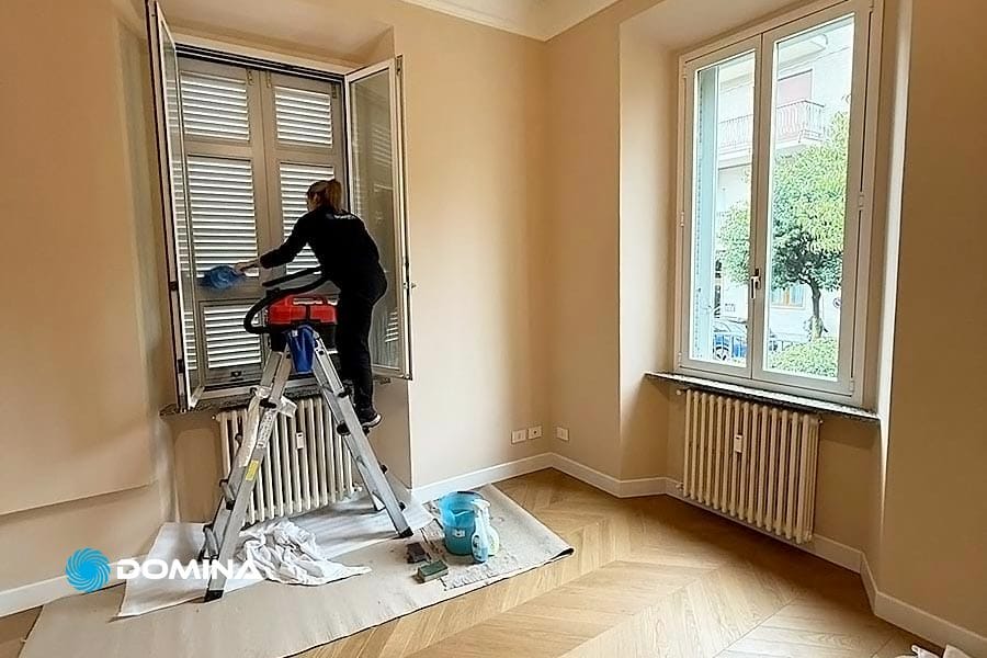 Nel cuore di Cassano Magnago, una persona è in piedi su una scala e pulisce le persiane delle finestre in una stanza non arredata con pavimento in parquet. Nelle vicinanze, un secchio e prodotti per la pulizia di Domina Pulizie lasciano intuire l'accurata pulizia post-ristrutturazione in corso.