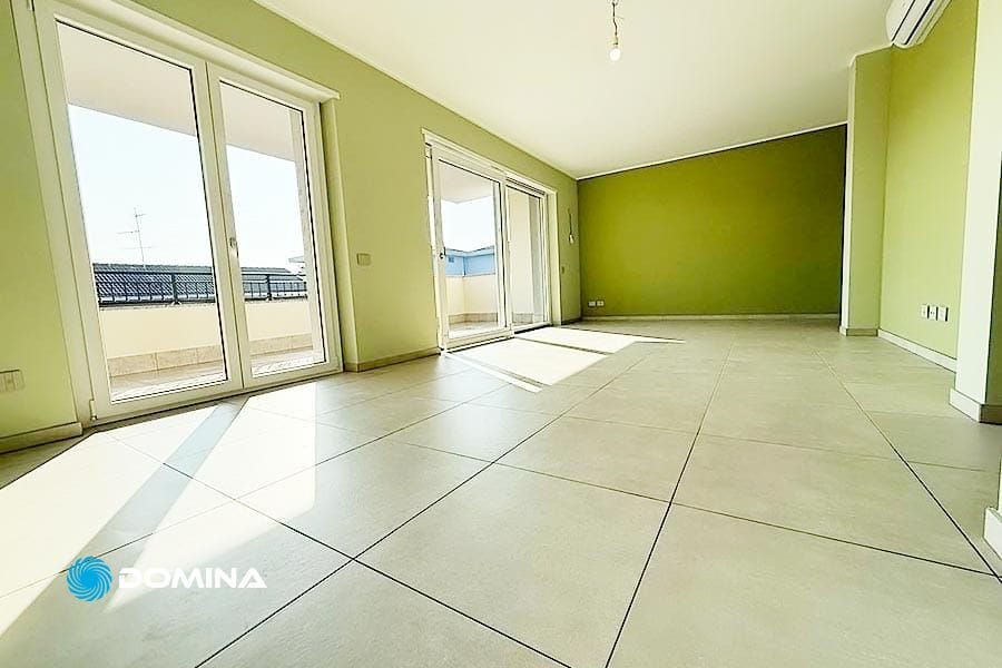 Ampia stanza vuota con grandi finestre e pareti verdi a Mariano Comense. La luce del sole filtra sul pavimento piastrellato beige, evidenziando la sensazione di spazio aperto. Un condizionatore è visibile sulla parete, perfettamente mantenuto da Domina Pulizie per una finitura impeccabile.