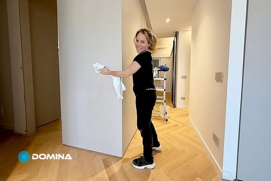 Una donna sorride mentre pulisce una parete bianca in un luminoso corridoio con pavimenti in legno, che espone la ditta pulizie post ristrutturazione Milano.