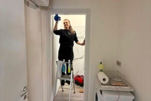 Persona in piedi su una scala a pioli che pulisce una porta a vetri in una piccola e luminosa stanza a Milano, vicino a Porta Genova, con prodotti per la pulizia a portata di mano: ideale per pulizie post ristrutturazione.