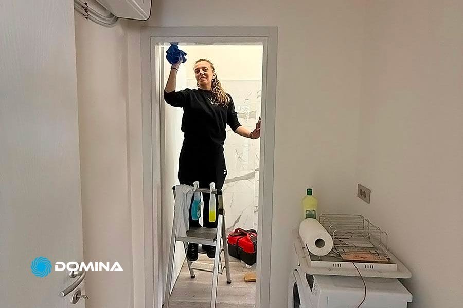 Persona in piedi su una scala a pioli che pulisce una porta a vetri in una piccola e luminosa stanza a Milano, vicino a Porta Genova, con prodotti per la pulizia a portata di mano: ideale per pulizie post ristrutturazione.