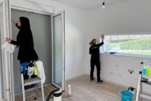 Due donne puliscono le finestre in una stanza luminosa e moderna con prodotti per la pulizia sul pavimento, specializzate in Pulizie Post Cantiere Como.