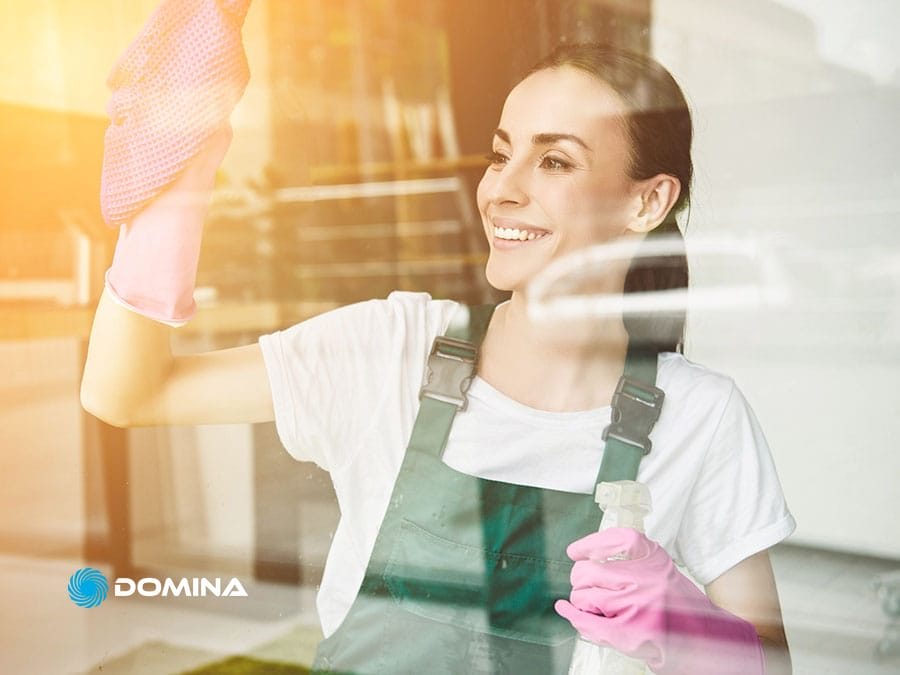 pulizie-uffici-5 Una donna sorridente con i guanti pulisce una finestra con un panno e uno spruzzino in una giornata di sole, a dimostrazione della cura e della precisione di Pulizie Uffici Garbagnate Milanese.