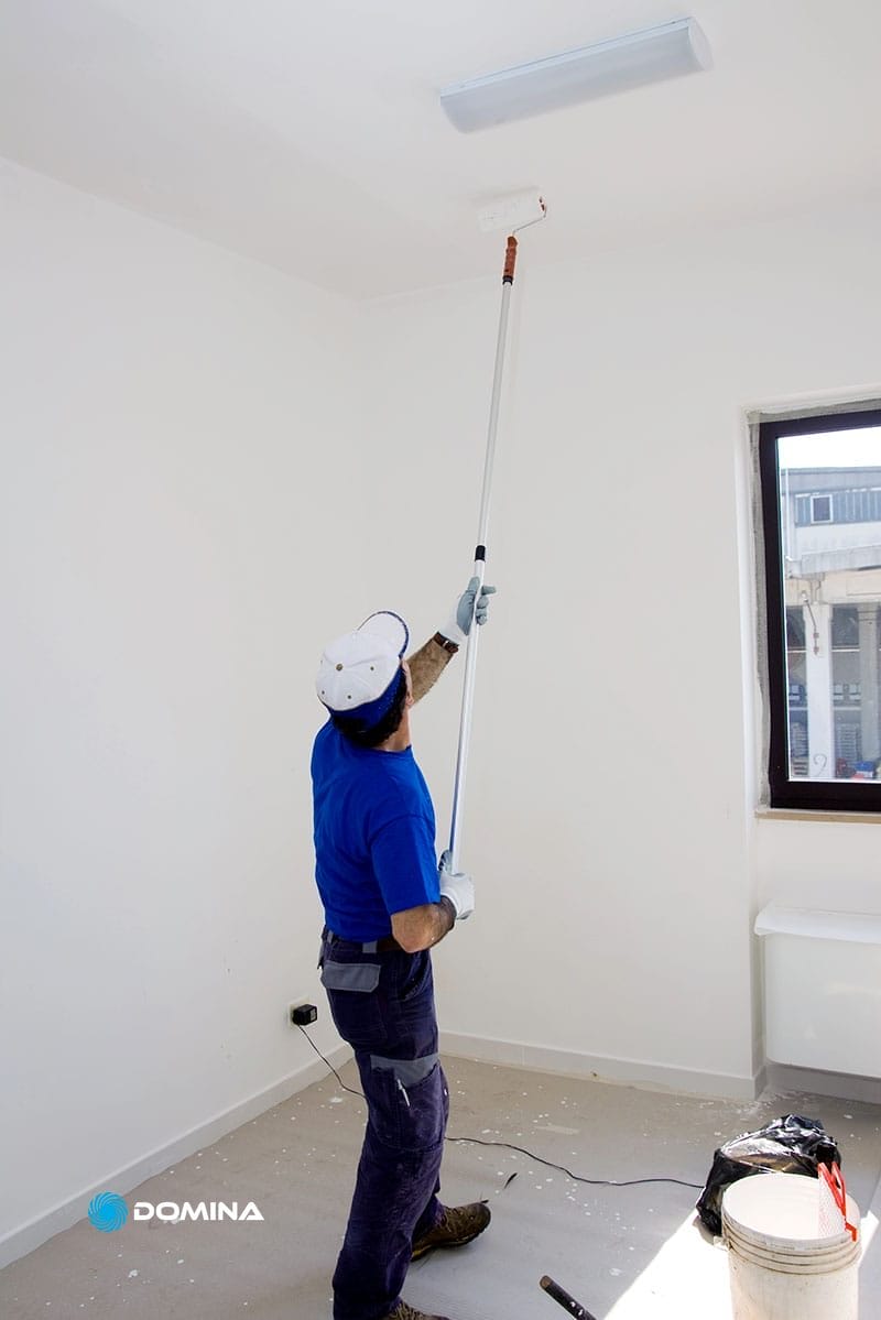 Una persona con una camicia blu dipinge un soffitto bianco con un rullo in una stanza luminosa e vuota, presentando servizi di imbiancatura professionale.