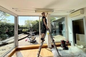 Persona in piedi su una scala in un interno di Cernobbio, che lavora al soffitto vicino a grandi finestre che si affacciano su un patio, gestendo Pulizie Primo Ingresso Appartamento con vista che ricorda il Lago di Como.