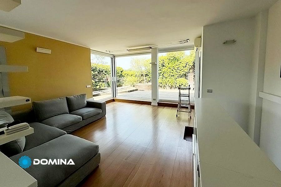 Soggiorno luminoso con divano grigio, pavimenti in legno, ampie finestre e vista su un patio soleggiato e sul giardino, perfettamente curato grazie a Pulizie Appartamenti Legnano.