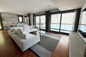 Soggiorno moderno con divano grigio, ampie finestre e vista sulla piscina; cucina aperta sullo sfondo, ideale per chi cerca pulizie villa di lusso Como.