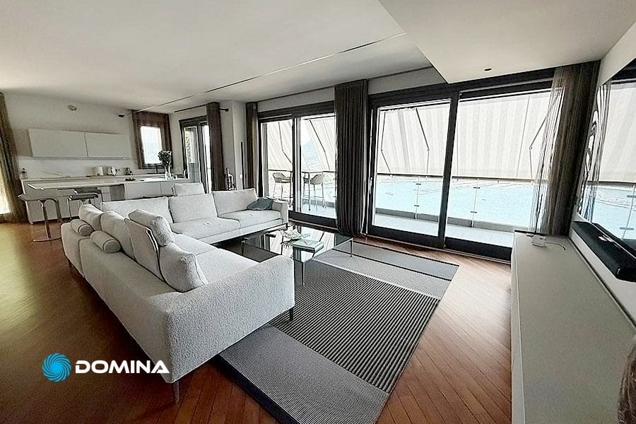 Soggiorno moderno con divano grigio, ampie finestre e vista sulla piscina; cucina aperta sullo sfondo, ideale per chi cerca pulizie villa di lusso Como.