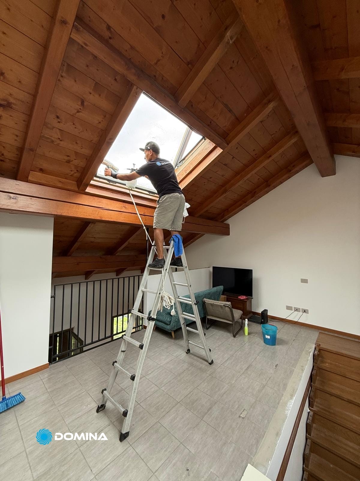 Persona in piedi su una scala che pulisce una finestra a lucernario in una Mansarda con Travi a Vista, che evidenzia Pulizie Post Cantiere.