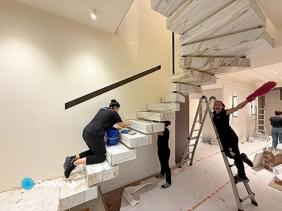 Tre persone puliscono una moderna scala di marmo in un appartamento di lusso a Milano; una saluta e sorride tenendo in mano uno spolverino rosa su una scala, mostrando la professionalità di un'impresa pulizie dopo pulizie post ristrutturazione.