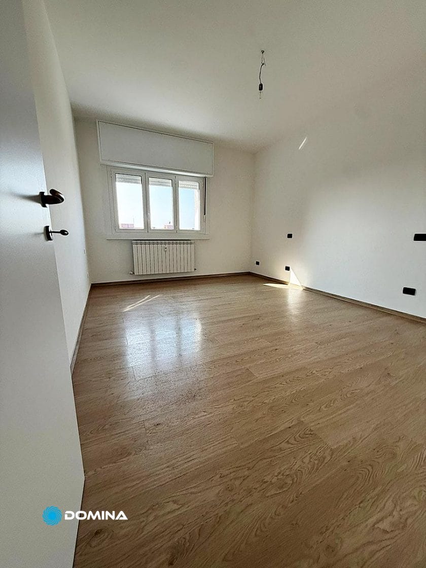 Una stanza luminosa e vuota con pavimento in legno chiaro, pareti bianche e una finestra che lascia entrare la luce naturale, ideale per le pulizie post cantiere Stazione Centrale Milano. Sotto la finestra c'è un radiatore e una plafoniera senza paralume.