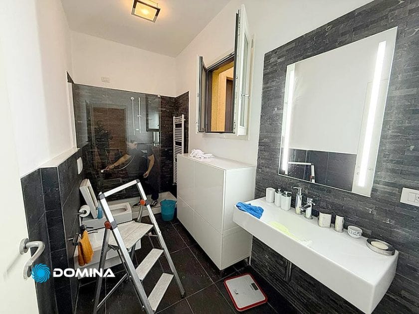 Un bagno moderno con pareti di piastrelle nere, una cabina doccia con porte in vetro e un lavandino bianco con un grande specchio illuminato: perfetto per chi cerca pulizie professionali Milano centro in appartamenti eleganti.