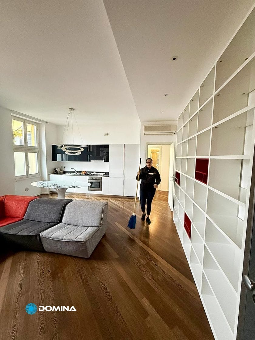 Una persona sta spazzando il pavimento in un soggiorno moderno e ordinato, con una scaffalatura bianca e un divano componibile grigio e rosso, che mostra i risultati di pulizie professionali Milano centro, con una cucina aperta e la luce naturale delle grandi finestre.