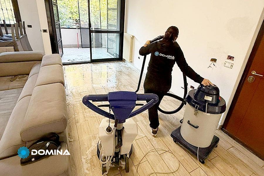 Una persona in abiti neri per le pulizie utilizza un aspirapolvere e una macchina per la pulizia per rimuovere l'acqua dal pavimento di un soggiorno moderno, ideale per le pulizie post cantiere Stazione Centrale Milano, con un divano grigio, grandi finestre e pavimento in legno bagnato.