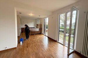Una persona passa lo straccio sul pavimento di legno di una stanza luminosa e non arredata, con grandi finestre e porte a vetri aperte su un giardino, probabilmente dopo Pulizie Post Ristrutturazione Varedo, con scatole e mobili coperti sullo sfondo.