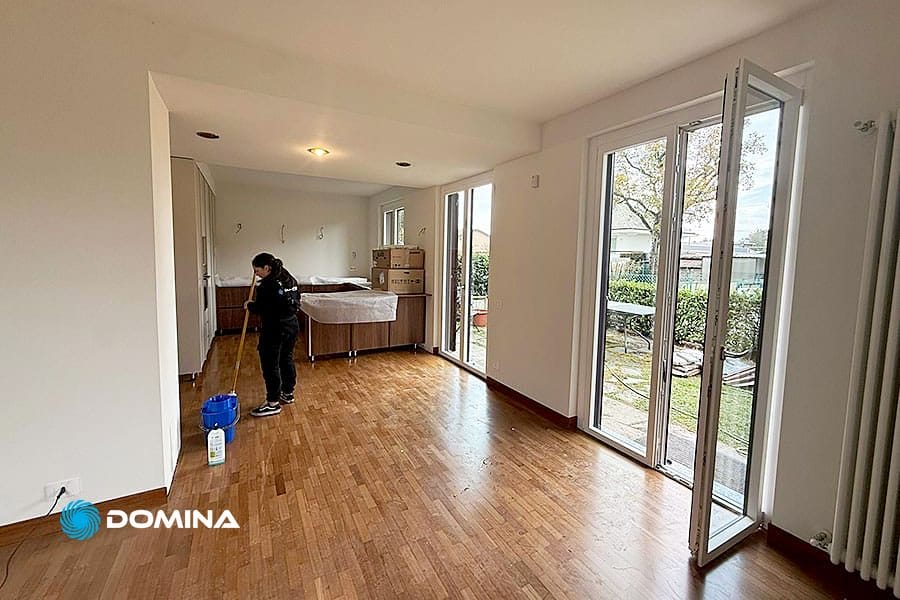 Una persona passa lo straccio sul pavimento di legno di una stanza luminosa e non arredata, con grandi finestre e porte a vetri aperte su un giardino, probabilmente dopo Pulizie Post Ristrutturazione Varedo, con scatole e mobili coperti sullo sfondo.
