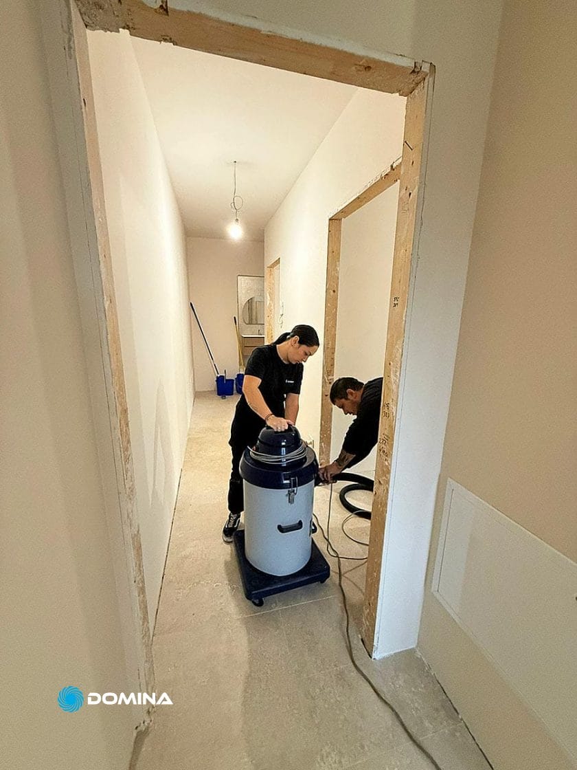 Due persone di Pulizie Fine Cantiere puliscono un corridoio luminoso in fase di ristrutturazione; una aziona un grande aspirapolvere mentre l'altra ne maneggia il tubo. Pareti grezze e materiale di pulizia arricchiscono l'atmosfera di Casa Pronta a Rho.