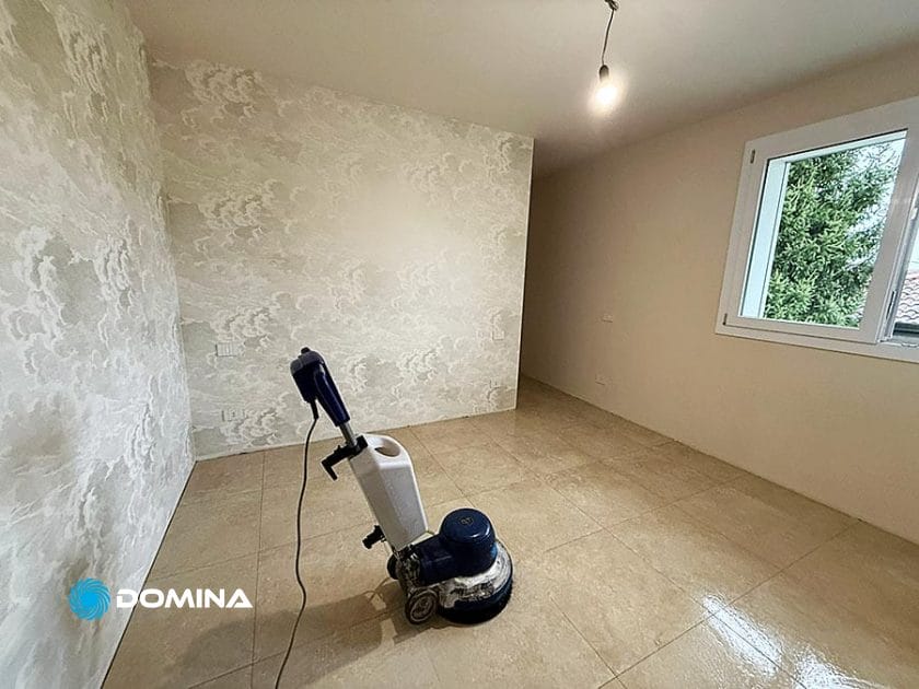 Una macchina lavapavimenti poggia su un pavimento piastrellato beige in una stanza vuota di Casa Pronta a Rho, con carta da parati a motivi su una parete, una sola lampadina appesa al soffitto e una finestra che mostra il verde all'esterno.