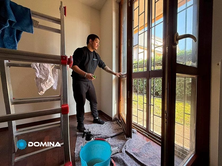 Una persona pulisce una grande porta a vetri con un panno, accanto a una scala di metallo e a un secchio blu su un pavimento ricoperto di asciugamani: la luce del sole entra e rivela il verde esterno. Pulizie Straordinarie Appartamento a Lainate in corso per risultati scintillanti.