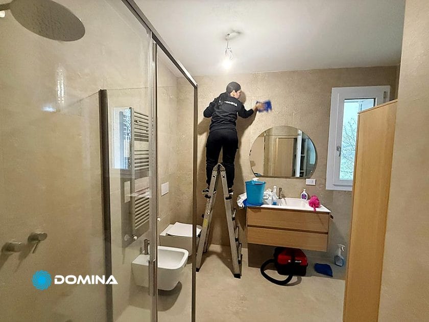 Una persona in piedi su una scaletta pulisce la parte superiore della parete del bagno vicino a una lampada. Il team di Casa Pronta è specializzato in Pulizie Fine Cantiere a Rho, assicurandosi che ogni angolo, dalla doccia in vetro al lavabo, sia immacolato.