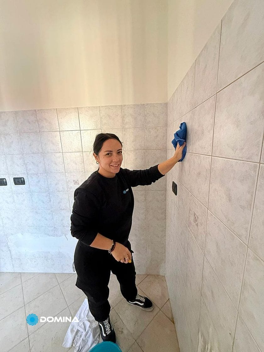Una donna vestita di nero sorride mentre pulisce le pareti piastrellate bianche con un panno blu in una stanza luminosa e vuota, illustrando Pulizie Straordinarie Appartamento a Lainate. Un secchio blu e un panno bianco sono appoggiati sul pavimento accanto a lei.