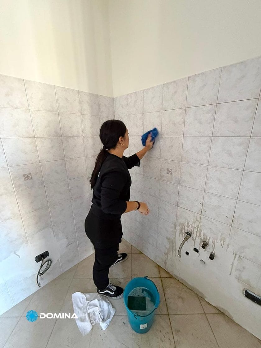 Una persona con i capelli scuri, vestita di nero, pulisce una parete piastrellata con un panno blu, probabilmente parte di Pulizie Straordinarie Appartamento a Lainate. Un secchio blu e un panno bianco alludono a una pulizia profonda o a una ristrutturazione, con macchie sul muro e buchi visibili in basso.