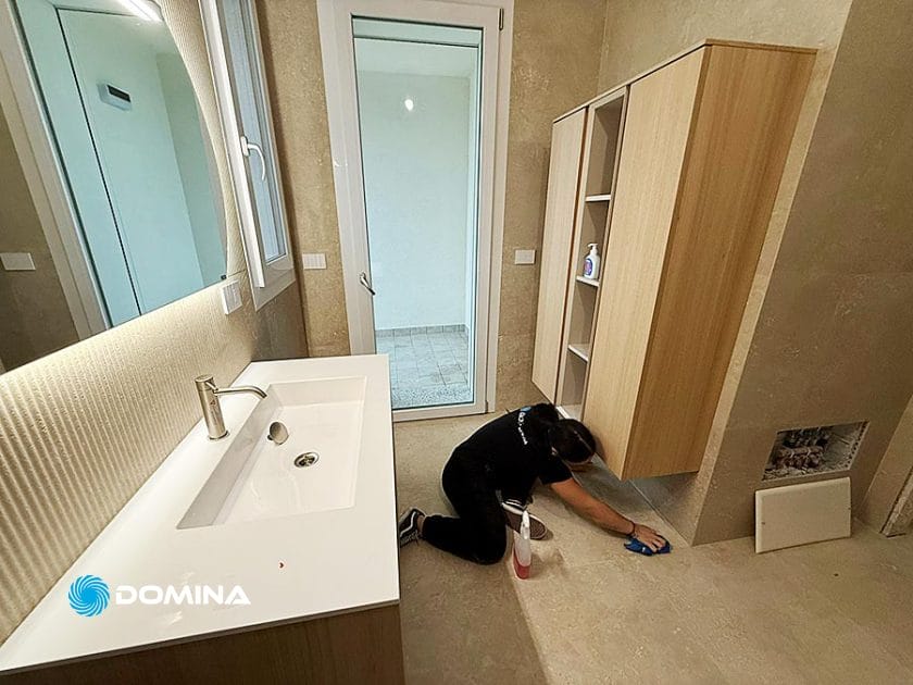 Una persona è inginocchiata sul pavimento di un bagno moderno, mentre pulisce vicino a un armadietto di legno, tipico di Pulizie Fine Cantiere a Rho. Lo spazio è caratterizzato da un lavandino bianco, uno specchio, pareti beige e una porta di vetro che conduce a una piccola area piastrellata.