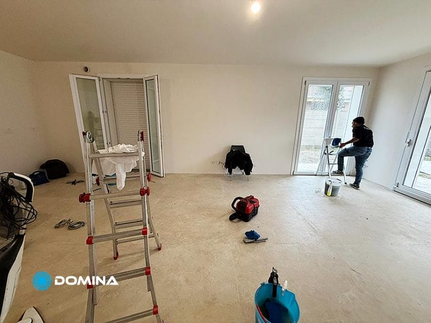 Una stanza per lo più vuota con pareti bianche e luce naturale proveniente da grandi finestre a Rho. Una persona si trova vicino a una finestra e lavora con un secchio di vernice e una scala a pioli, perfetta per le Pulizie Fine Cantiere prima di consegnare una Casa Pronta.