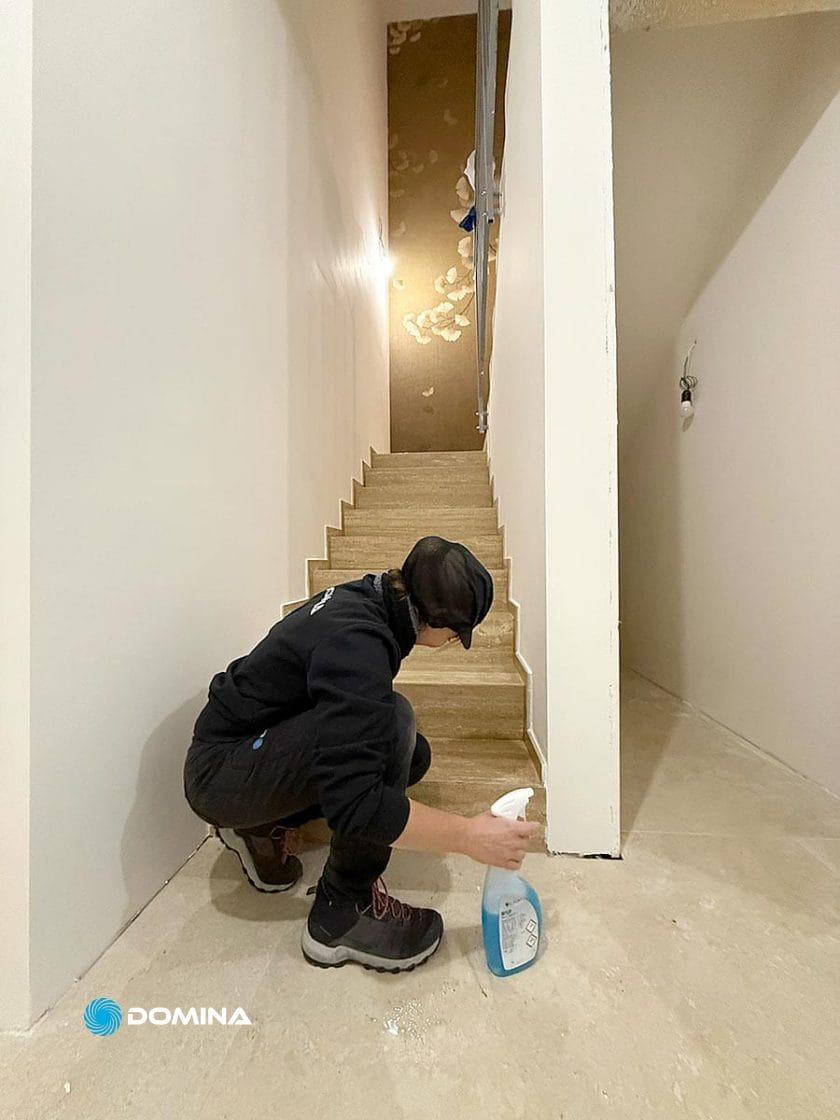 Una persona con felpa e berretto nero si inginocchia su un pavimento nudo, pulendo la base di una scala con una bomboletta spray-Pulizie Fine Cantiere in corso per realizzare la vostra Casa Pronta a Rho dopo la costruzione o la ristrutturazione.