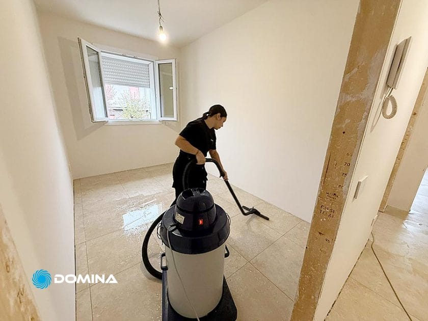 Una persona usa un grande aspirapolvere per pulire il pavimento piastrellato di una stanza vuota e appena dipinta a Rho, dove si svolgono le Pulizie Fine Cantiere per una Casa Pronta, con la luce del sole che filtra da una finestra aperta e una plafoniera spoglia in alto.