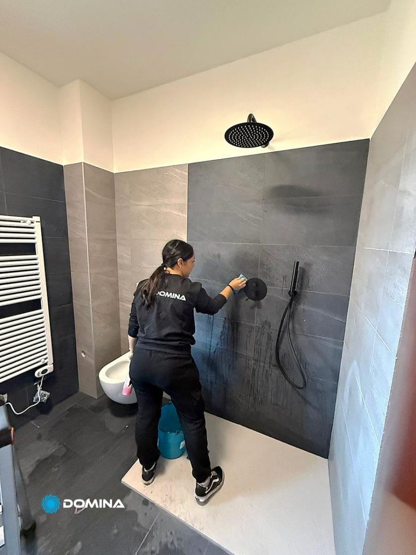 Una donna vestita di nero pulisce con una spugna la parete piastrellata di una doccia moderna in un bagno. Un secchio e dei prodotti per la pulizia sono nelle vicinanze - parte di Pulizie Straordinarie Appartamento a Lainate.