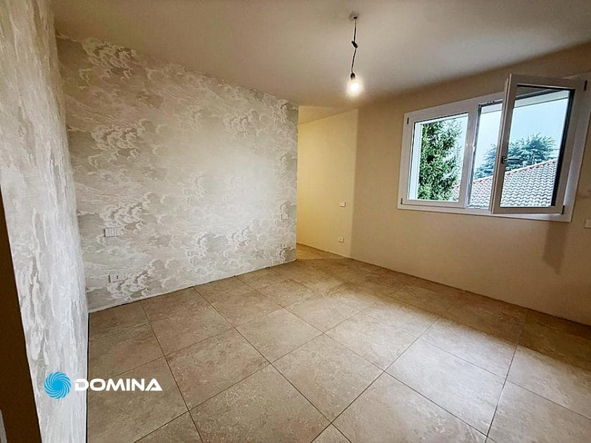 Una stanza vuota a Rho con pavimenti in piastrelle beige, una parete d'accento decorata con carta da parati, pareti color crema e una finestra aperta sugli alberi: perfetta per i servizi di Casa Pronta o Pulizie Fine Cantiere.