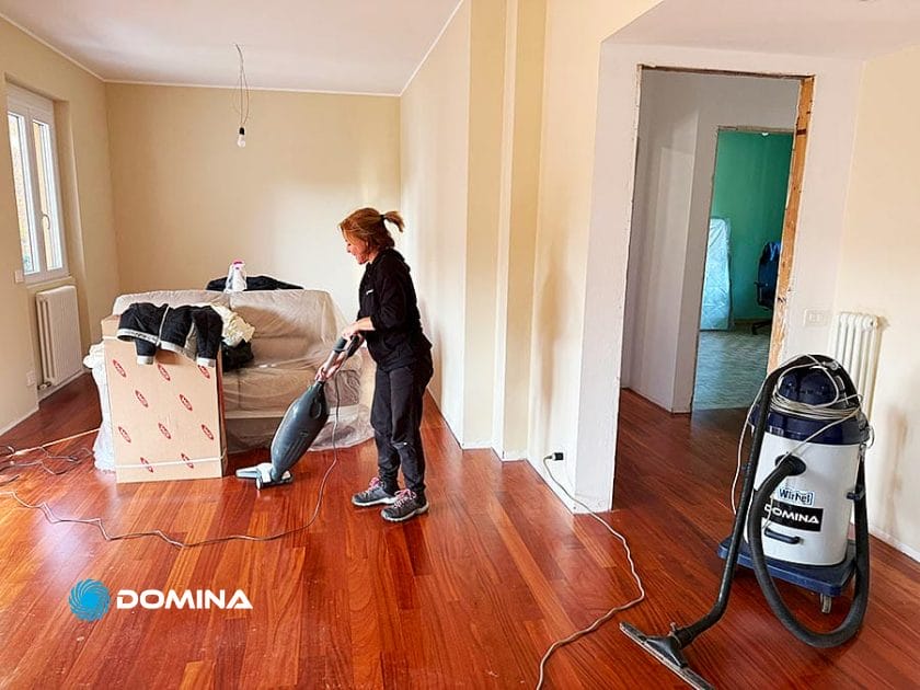 Una donna passa l'aspirapolvere sul pavimento in legno di una stanza parzialmente arredata durante Pulizie Straordinarie Appartamento a Lainate. Un divano coperto di coperte e scatoloni si trova accanto alla finestra, mentre alle pareti spoglie e chiare sono appesi gli attrezzi per la pulizia.
